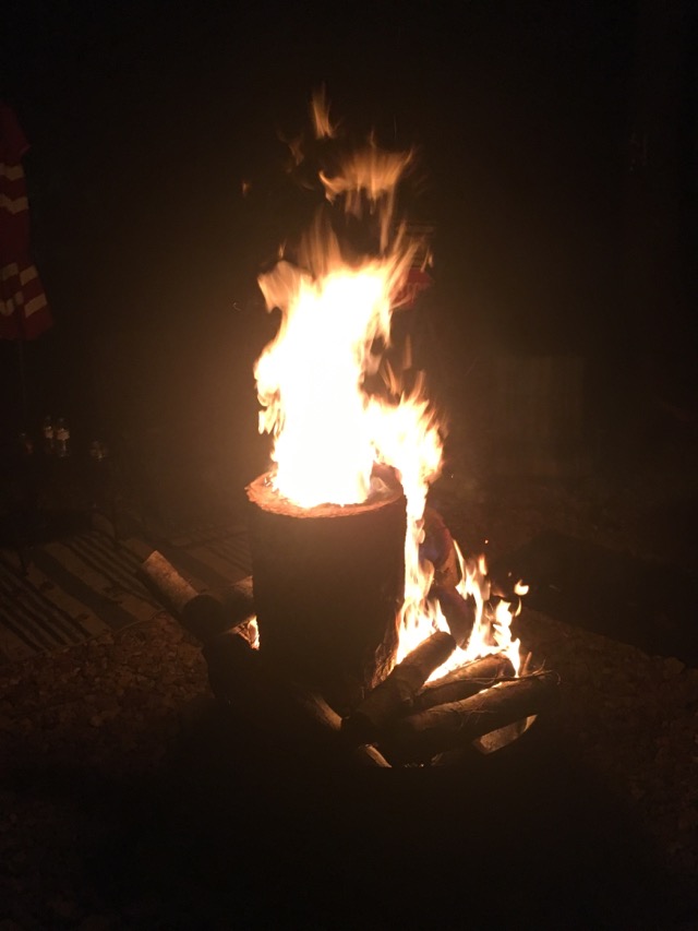 An outdoor campfire. It's dark and hard to see anything around the fire. To the left their appears to be a rug and umbrella just at the edge of the photo.